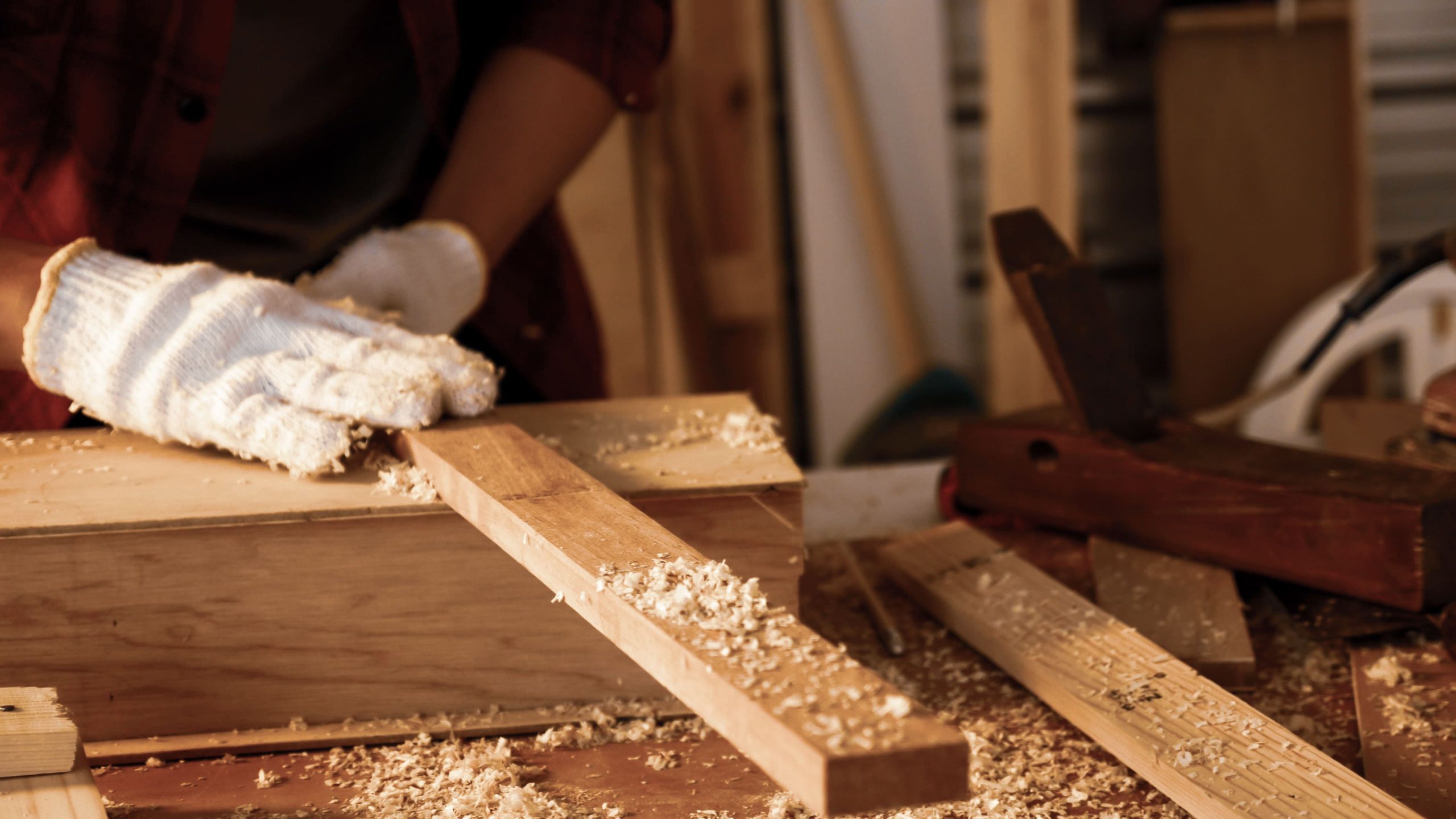 Person wearing white gloves planing a wooden board on a cluttered workbench covered with wood shavings.