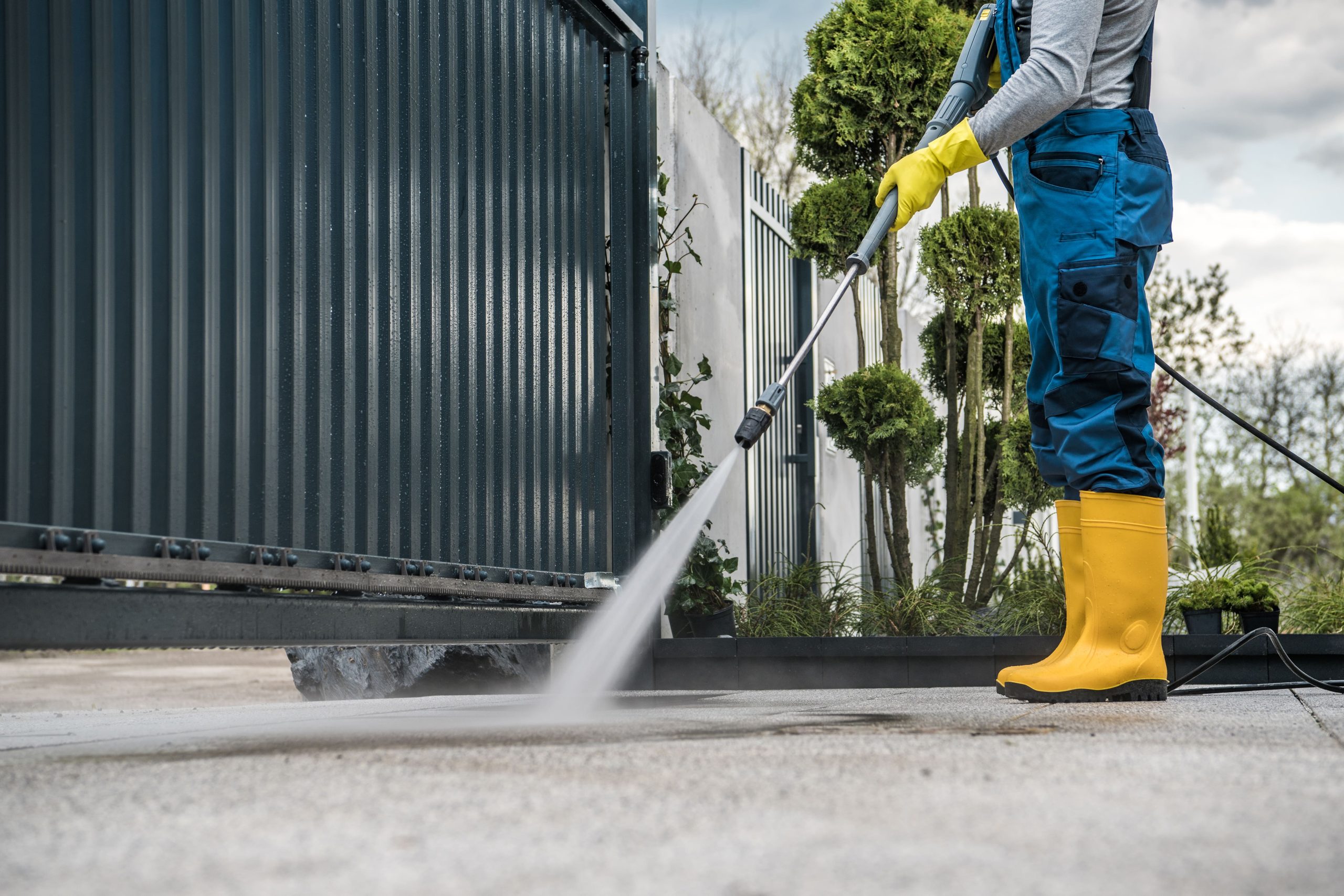 Person wearing yellow rubber boots and gloves pressure washing a driveway near a dark metal gate and green shrubs.