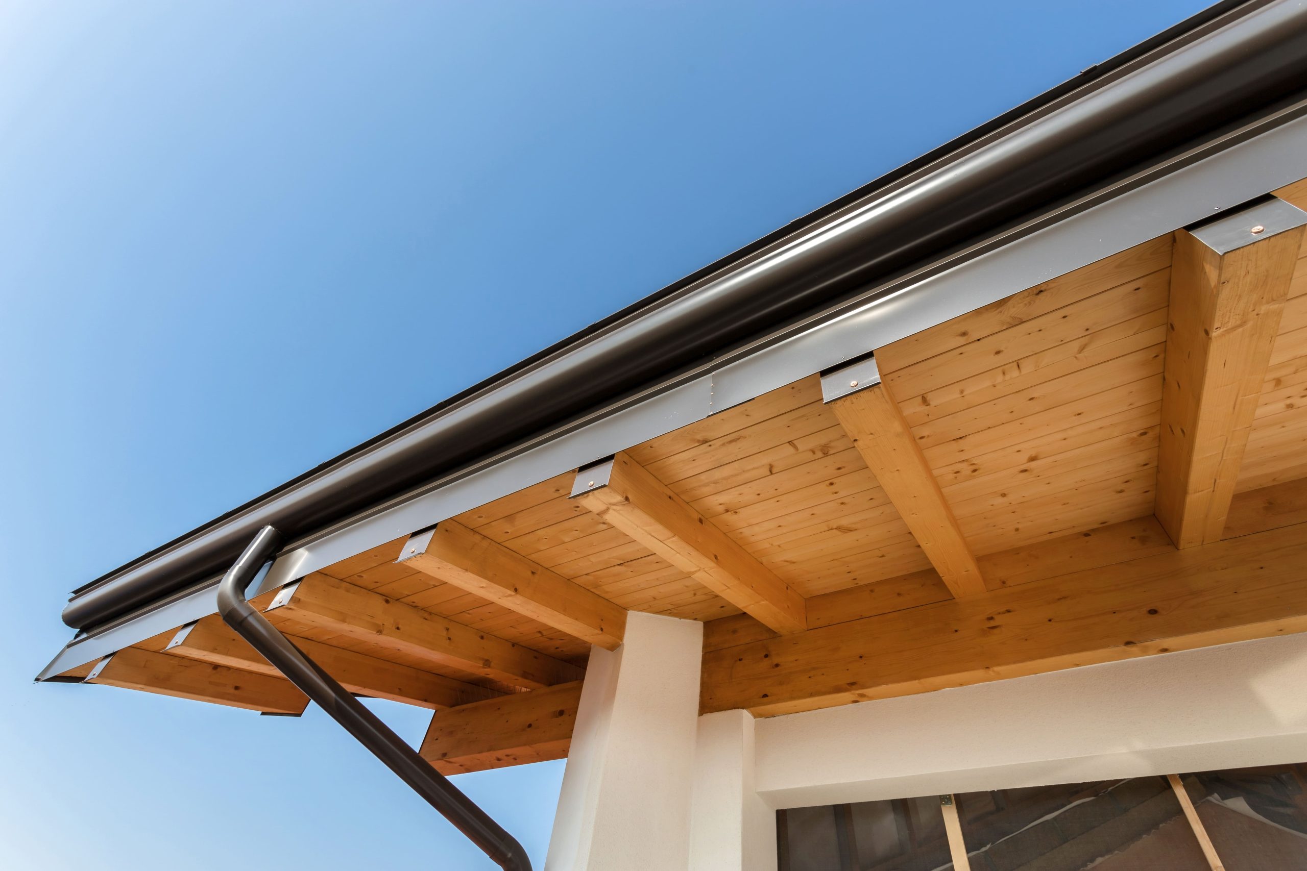Underneath view of a house roof with exposed wooden beams and a black gutter against a clear blue sky.