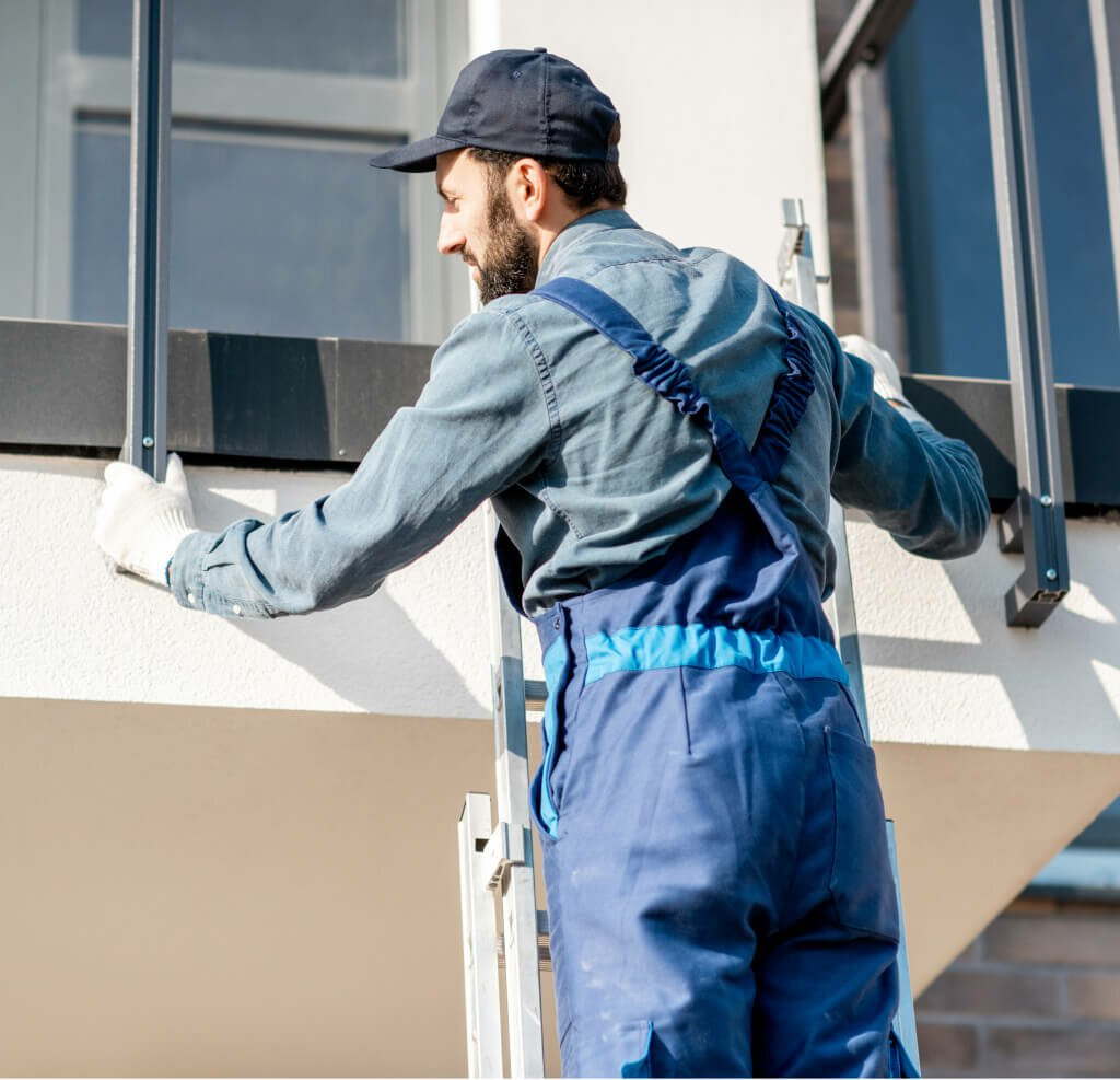 A worker in blue overalls and a cap is installing a railing on a balcony, using a ladder for support. The scene depicts a home improvement project focused on enhancing safety and aesthetics.